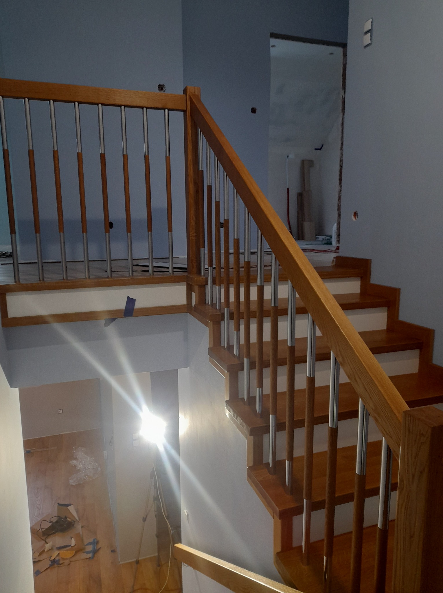 Nowoczesne schody z drewnianymi stopniami i balustradą z elementami drewnianymi i metalowymi, widok z góry, oświetlenie budowlane w tle, częściowo wykończone wnętrze.