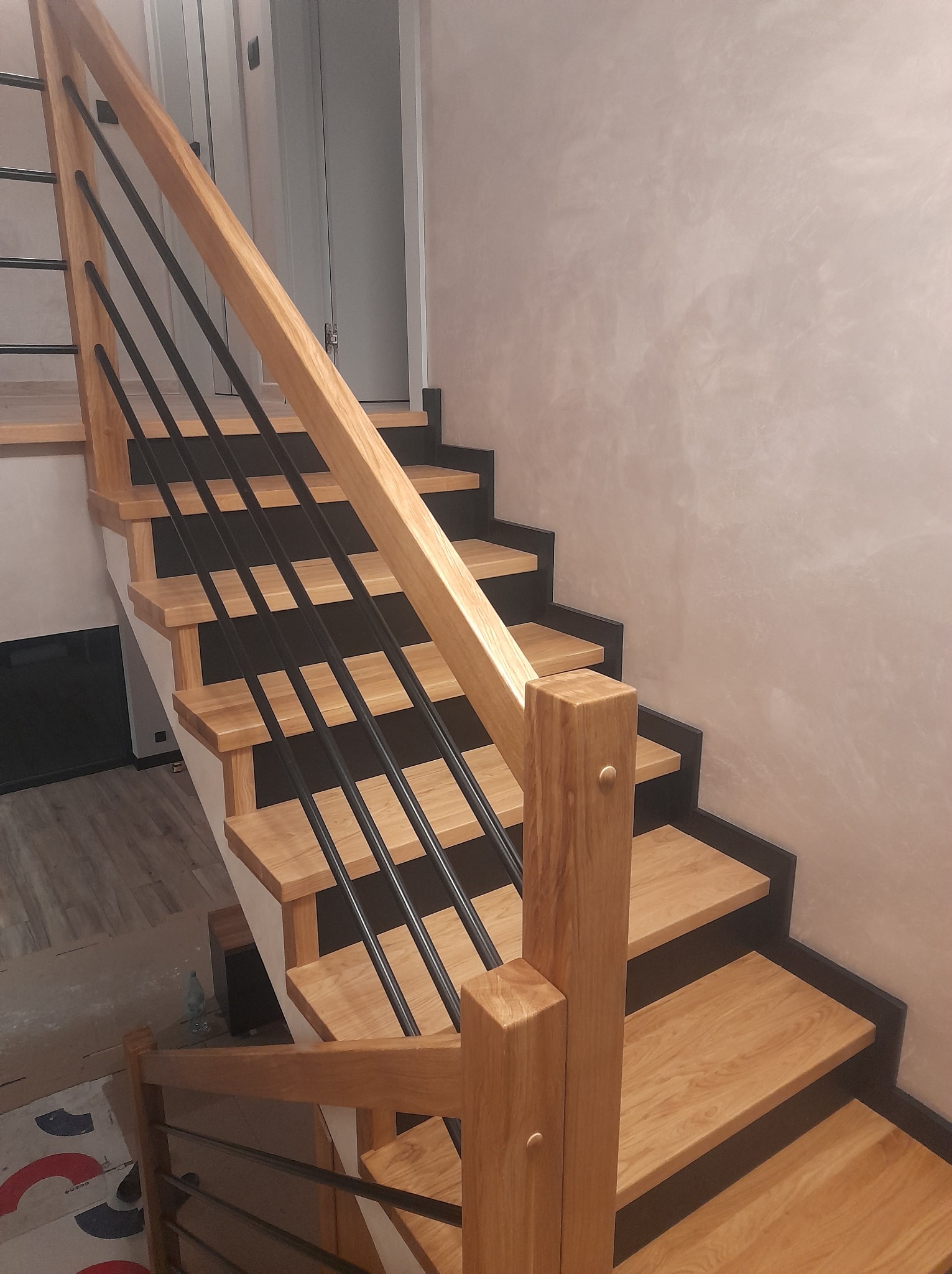 Nowoczesne schody z drewnianymi stopniami i czarnymi, metalowymi balustradami, widoczne z dołu, z elementami wykończenia w kolorze dębu.