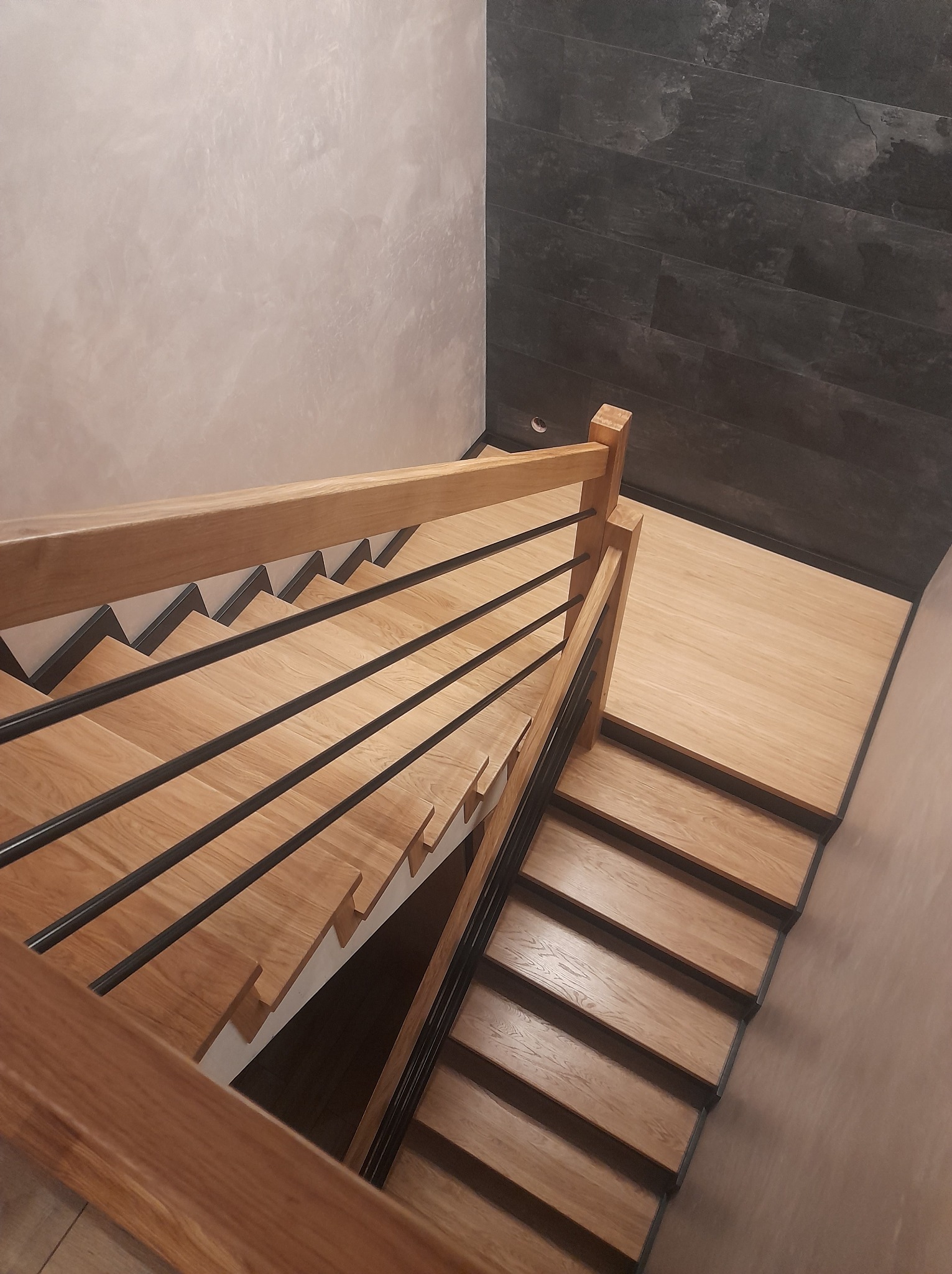 Nowoczesne schody z jasnego drewna z czarnymi, poziomymi balustradami, widok z góry, ściana wykończona w szarym kamieniu.