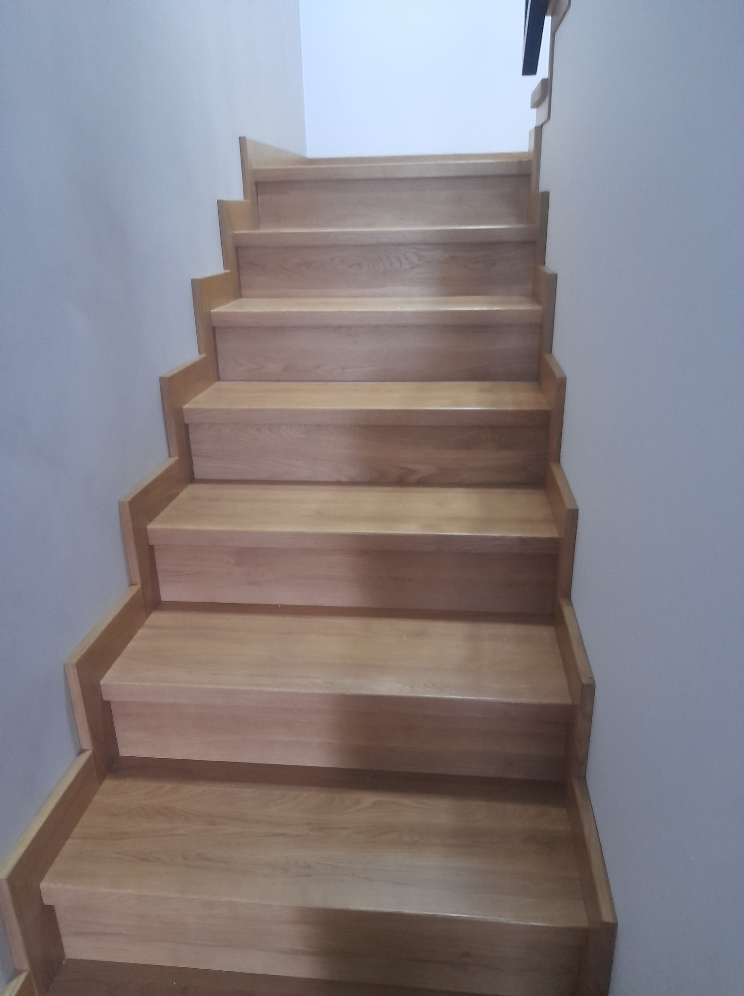 Proste, drewniane schody z jasnego drewna, bez balustrady, przy szarej ścianie, widok od dołu.