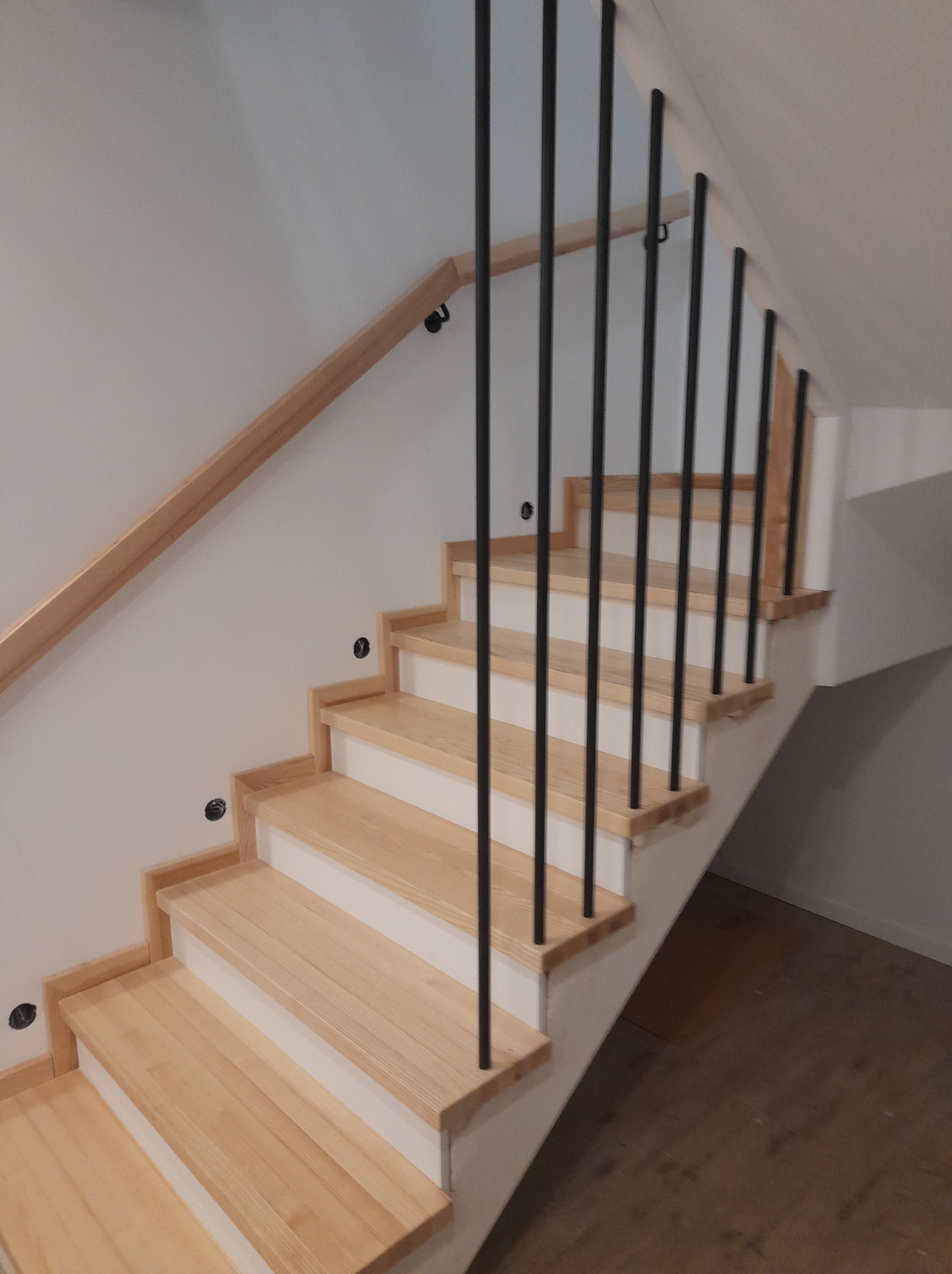 Jasne, drewniane stopnie schodów z czarnymi, metalowymi słupkami balustrady i drewnianą poręczą na białej ścianie, widoczne czarne zaślepki po otworach.