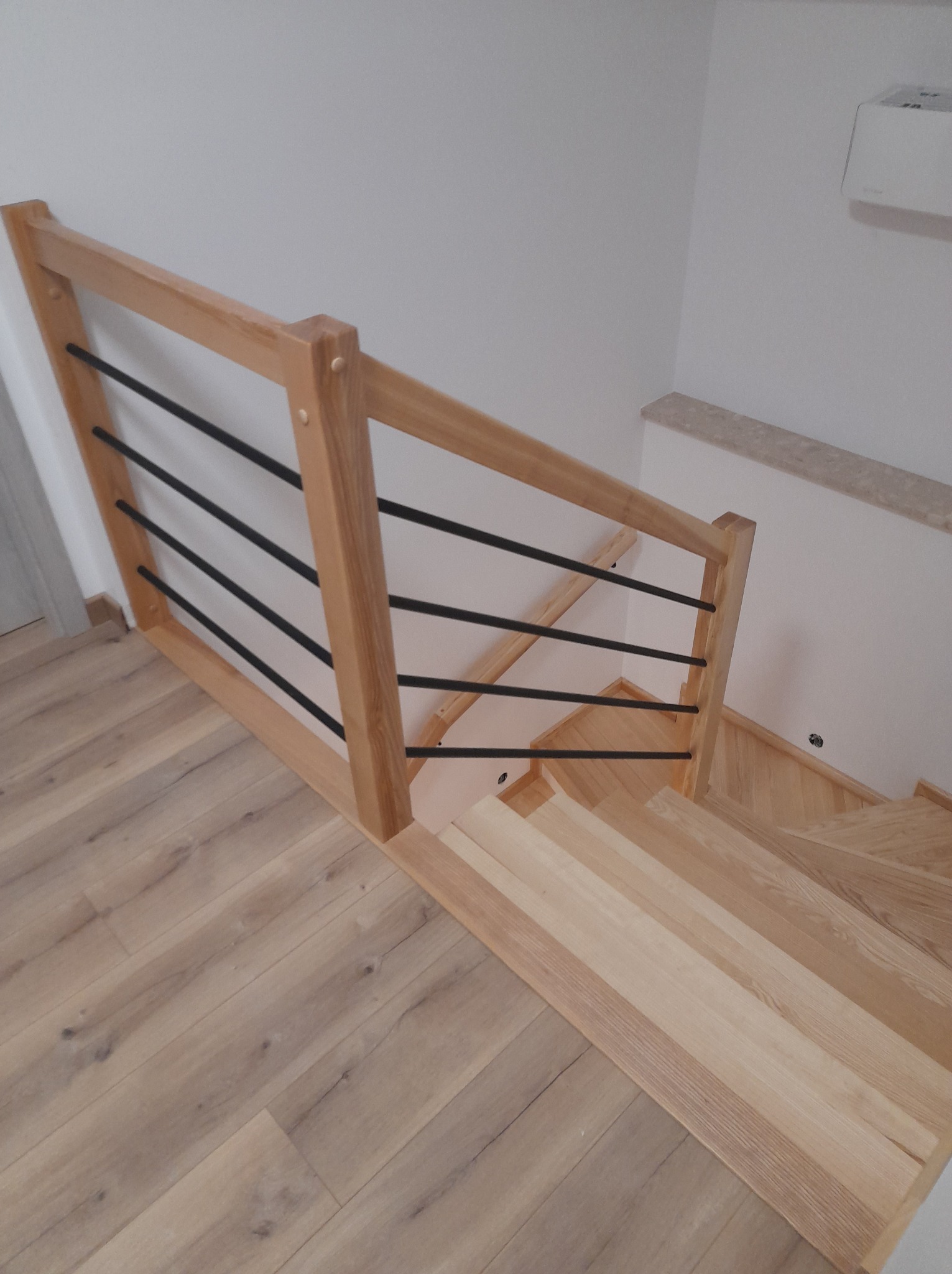 Jasne, drewniane schody z balustradą o minimalistycznym designie, gdzie drewniana rama łączy się z czarnymi, poziomymi prętami. Widok z góry, ukazujący naturalne usłojenie drewna i subtelne...