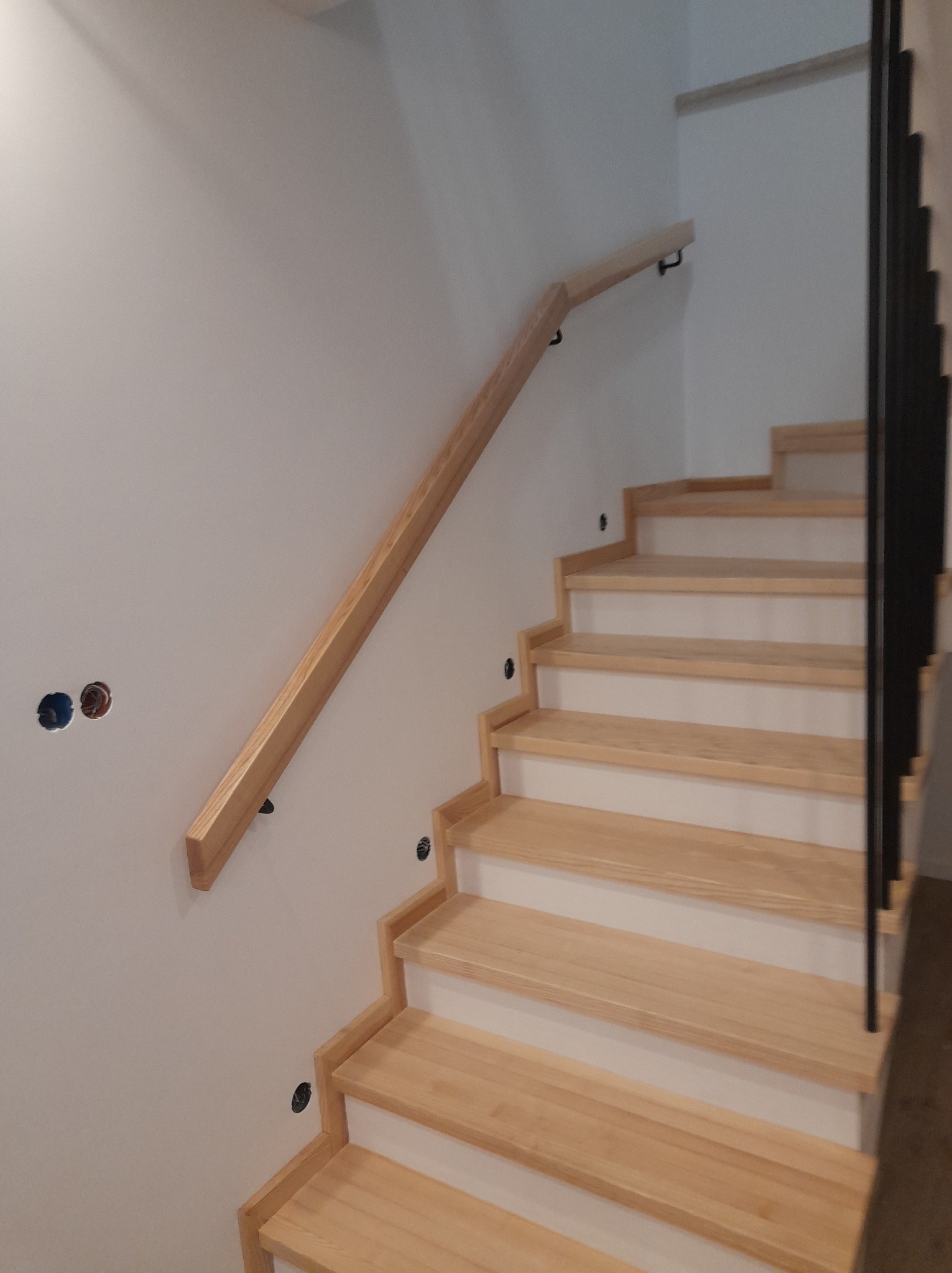 Jasne, drewniane stopnie schodów z minimalistyczną, drewnianą poręczą i czarną balustradą w domu w Straszowie, widoczne otwory na oświetlenie w ścianie.