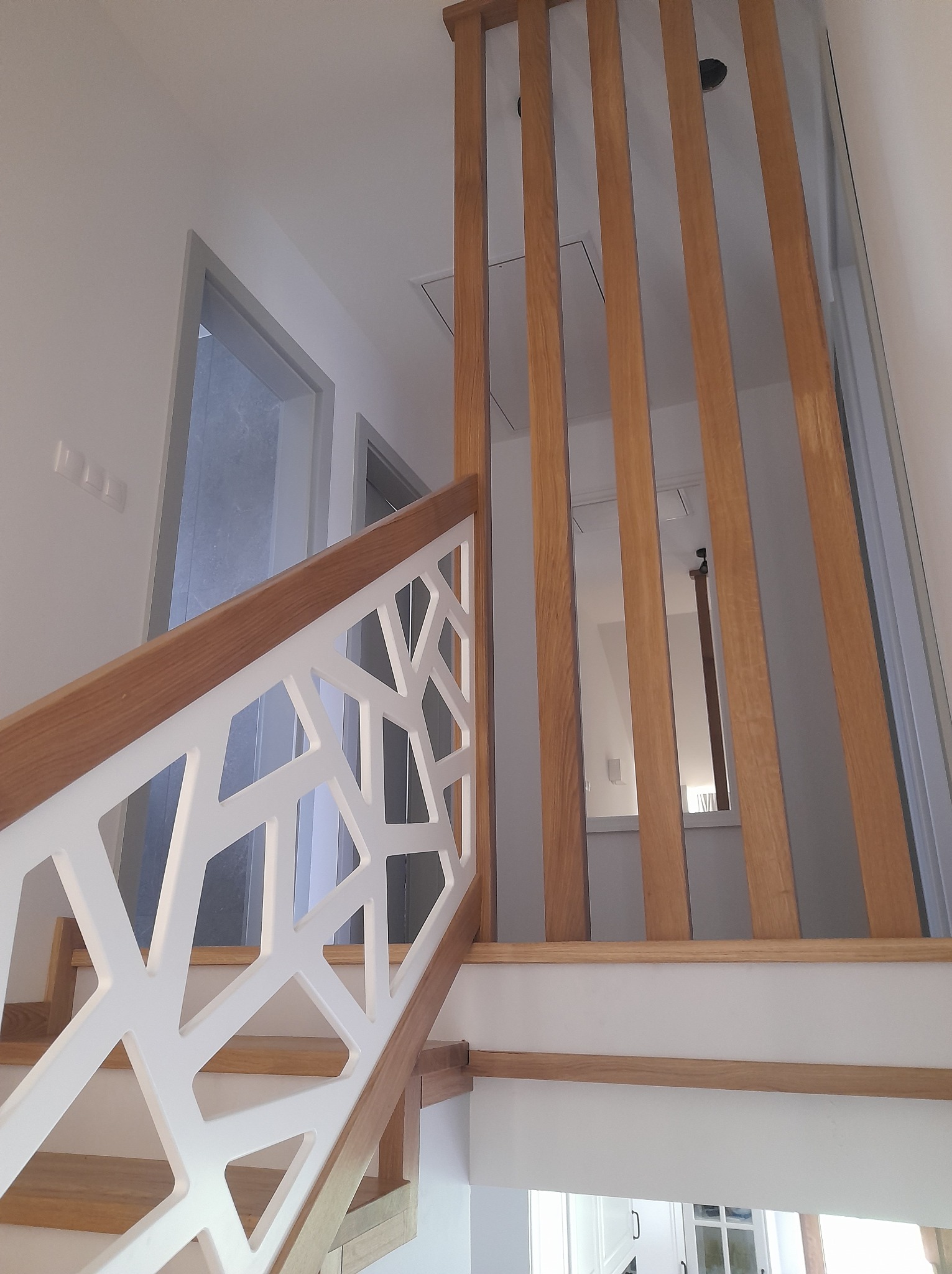 Nowoczesne schody z białą balustradą o geometrycznym wzorze i pionowymi drewnianymi lamelami, widok z dołu na górę.