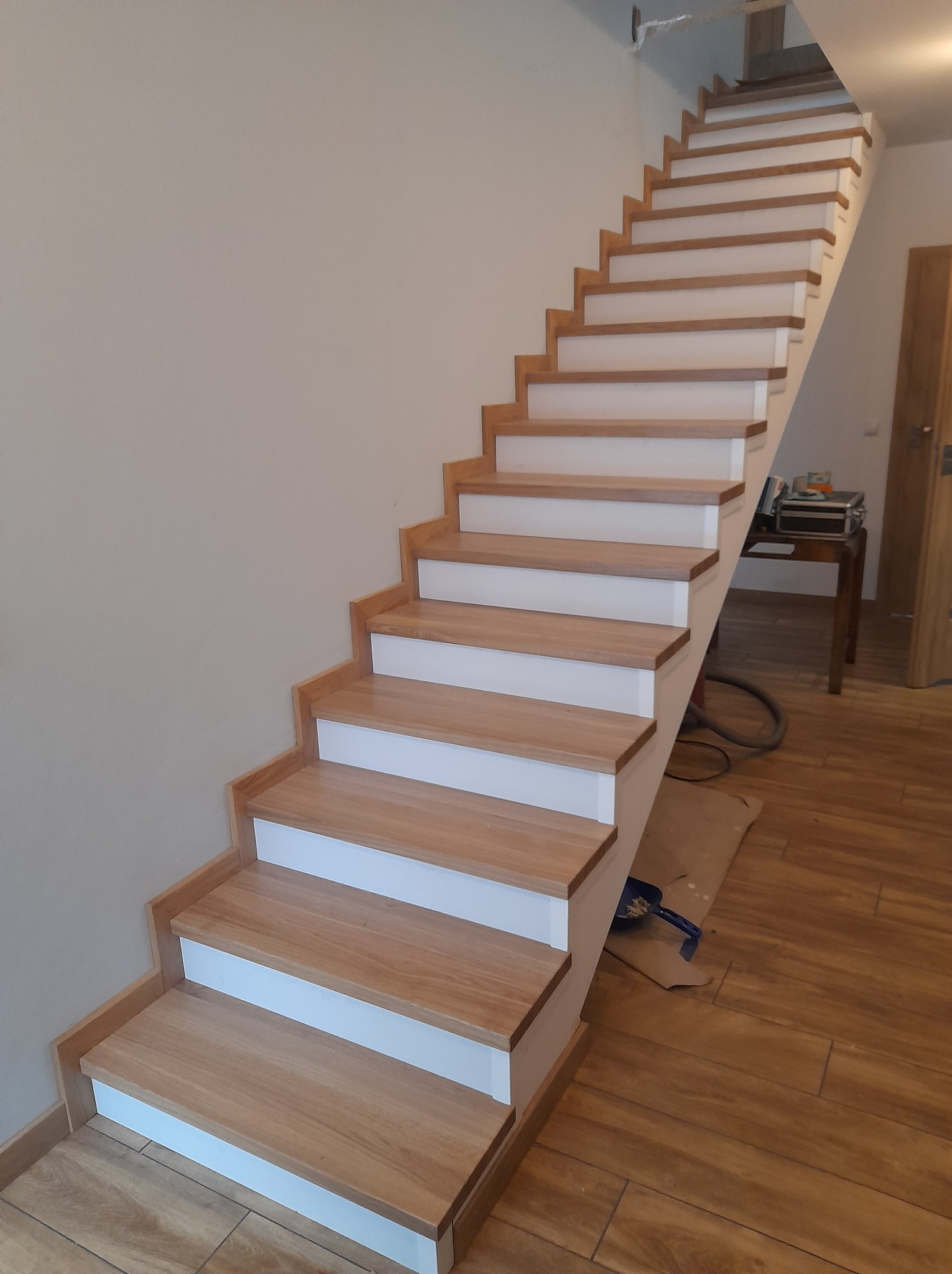 Nowoczesne schody z drewnianymi stopniami i białymi podstopnicami, widoczne narzędzia malarskie na podłodze, jasne wnętrze.
