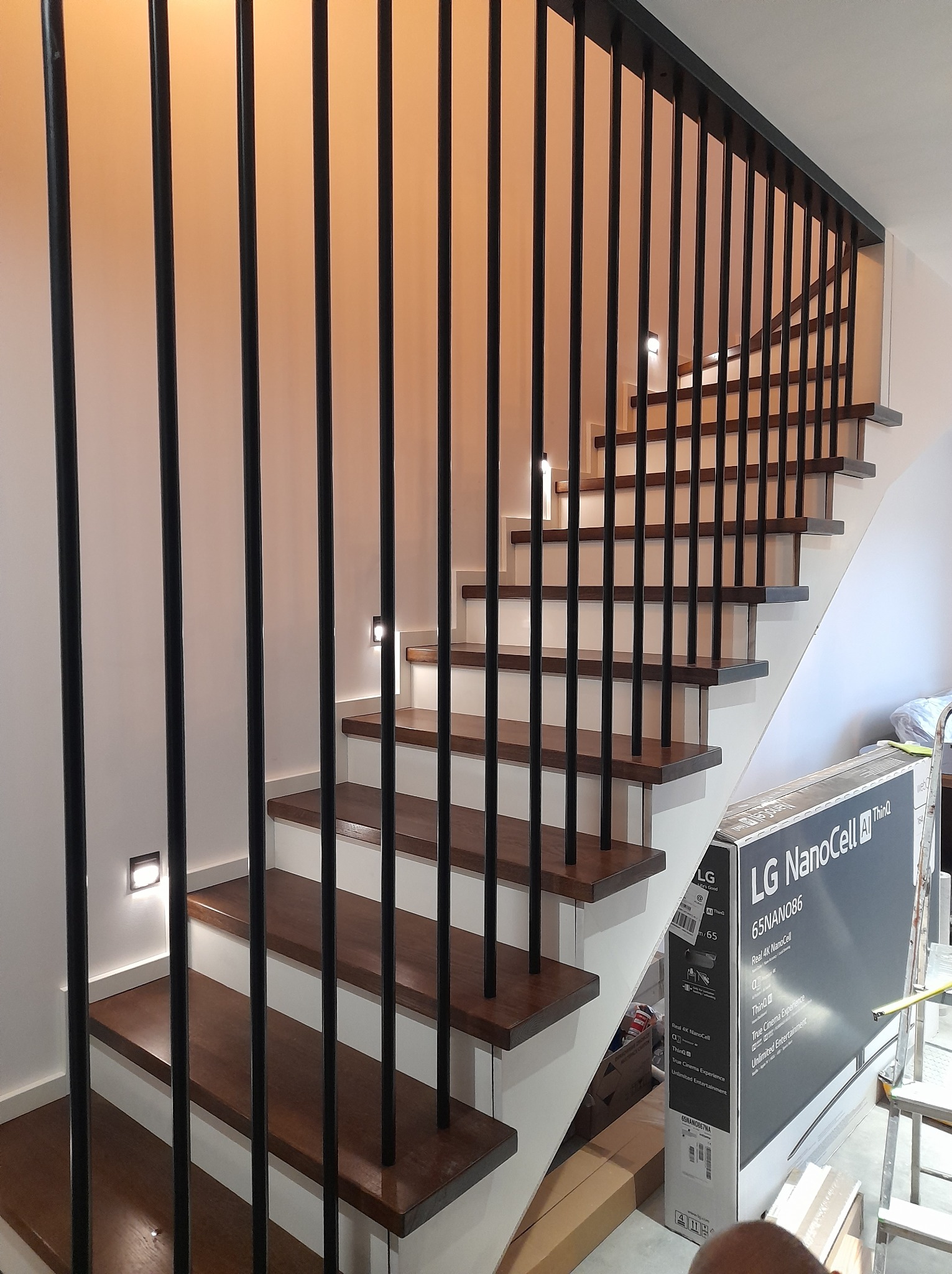 Nowoczesne schody drewniane z czarną balustradą z pionowych prętów, oświetleniem LED w ścianie, z widocznym kartonem po telewizorze LG NanoCell w tle.