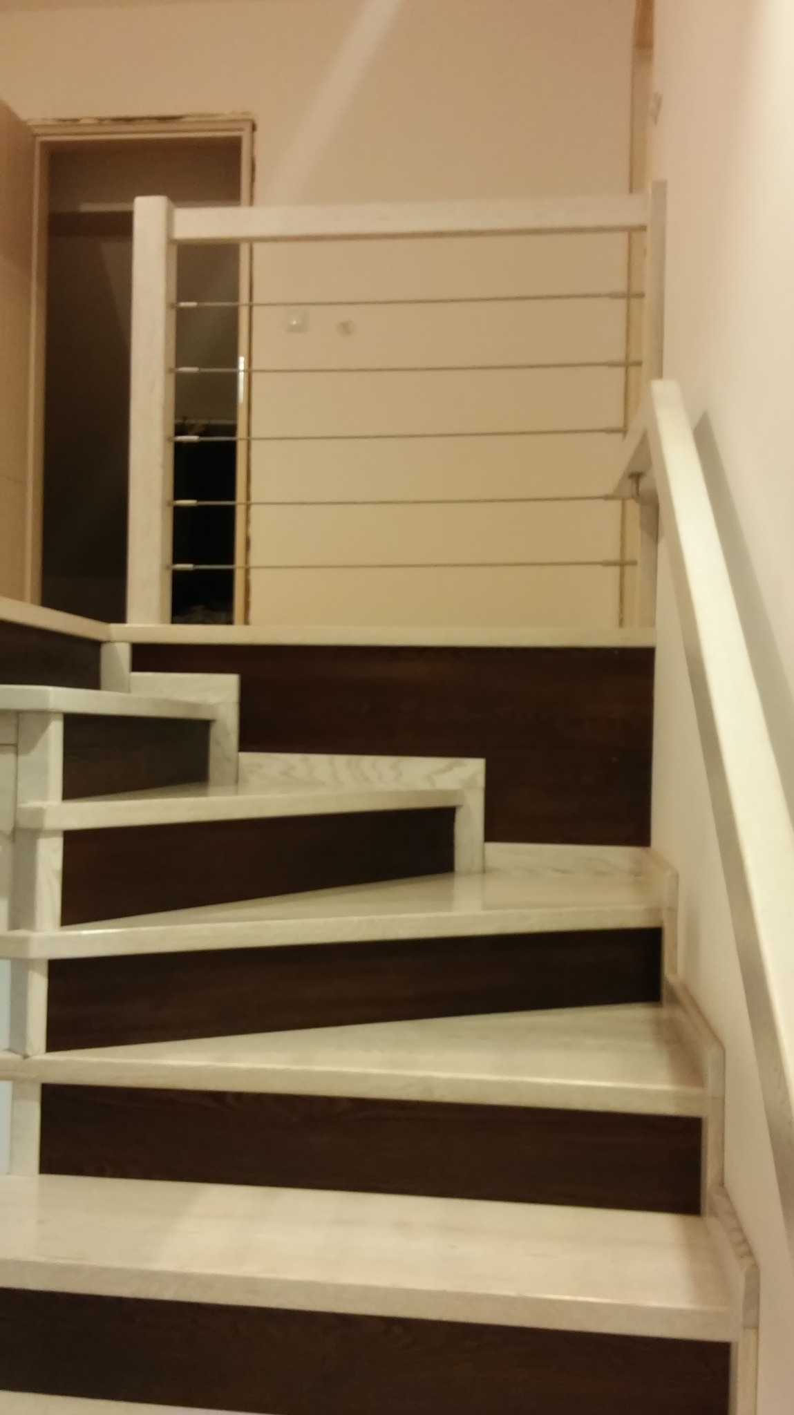 Nowoczesne schody z marmurowymi stopniami i ciemnobrązowymi podstopnicami, zabezpieczone balustradą z białych słupków i poziomych metalowych linek.