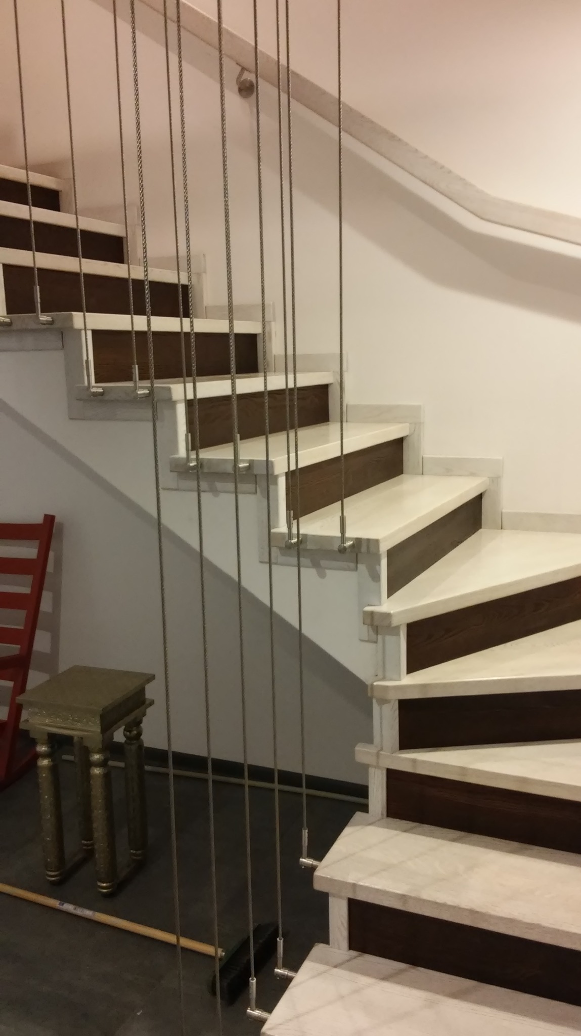 Spiralne schody z marmurowymi stopniami i ciemnobrązowymi podstopnicami, zabezpieczone metalowymi linkami, widoczne z dołu, z fragmentem czerwonego krzesła i złotego stołka w tle.