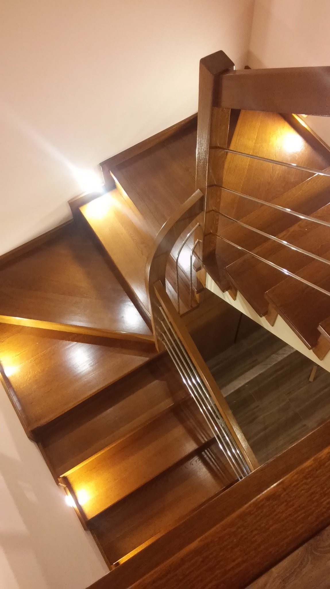 Klatka schodowa z drewna o ciepłym odcieniu, spiralne schody z balustradą z metalowych prętów, oświetlenie punktowe podkreśla fakturę drewna.