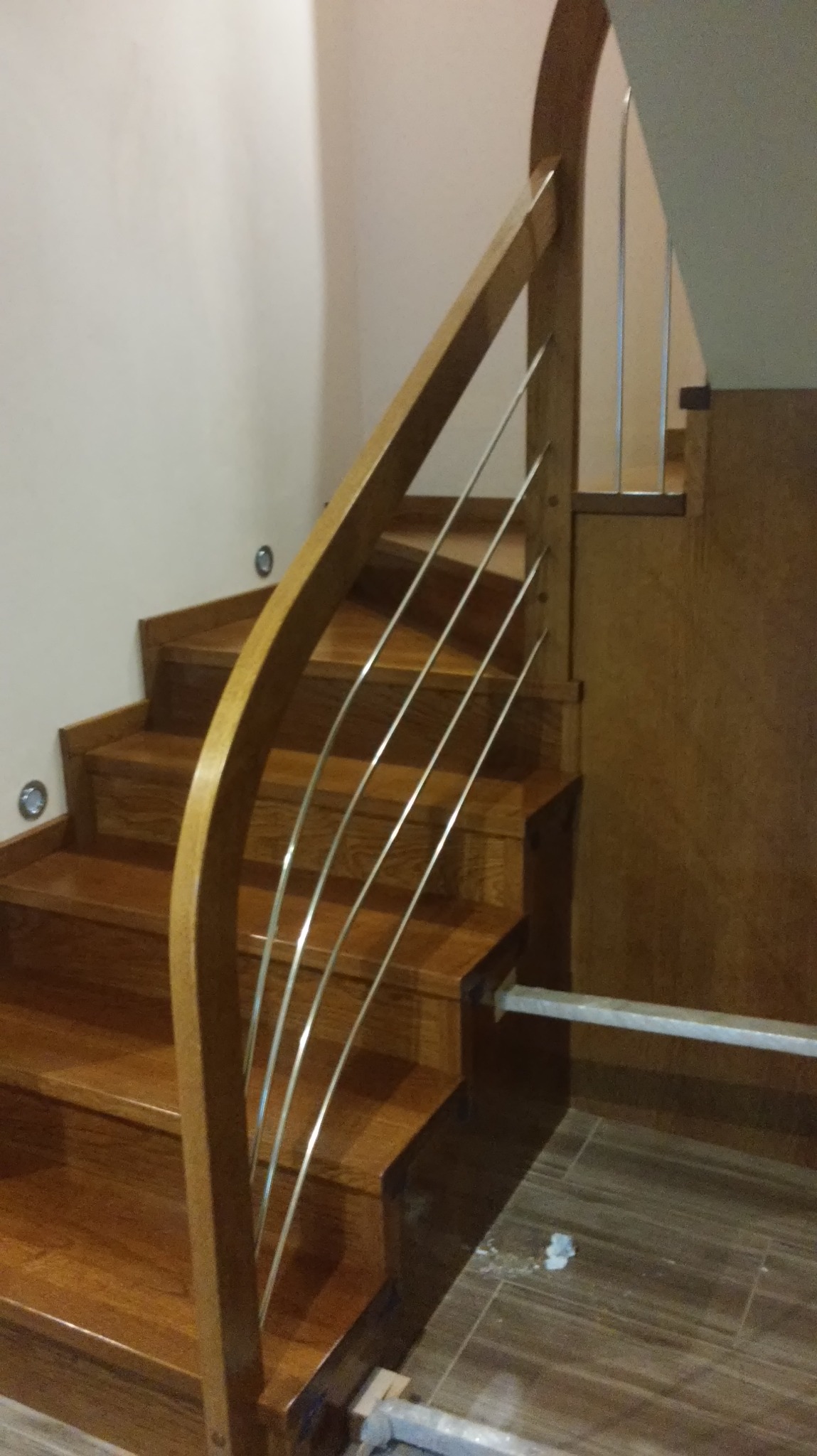 Klatka schodowa z drewnianymi stopniami i balustradą z giętego drewna oraz metalowymi prętami, widoczne oświetlenie punktowe na ścianie i fragment podłogi z zabrudzeniami po montażu.