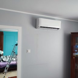 OPTIMA AGNIESZKA CZYŻYCKA - Biały klimatyzator ścienny zamontowany w szarym pokoju, widoczna drabina w przejściu do pokoju z turkusowymi ścianami, obok stoi ciemna witryna.