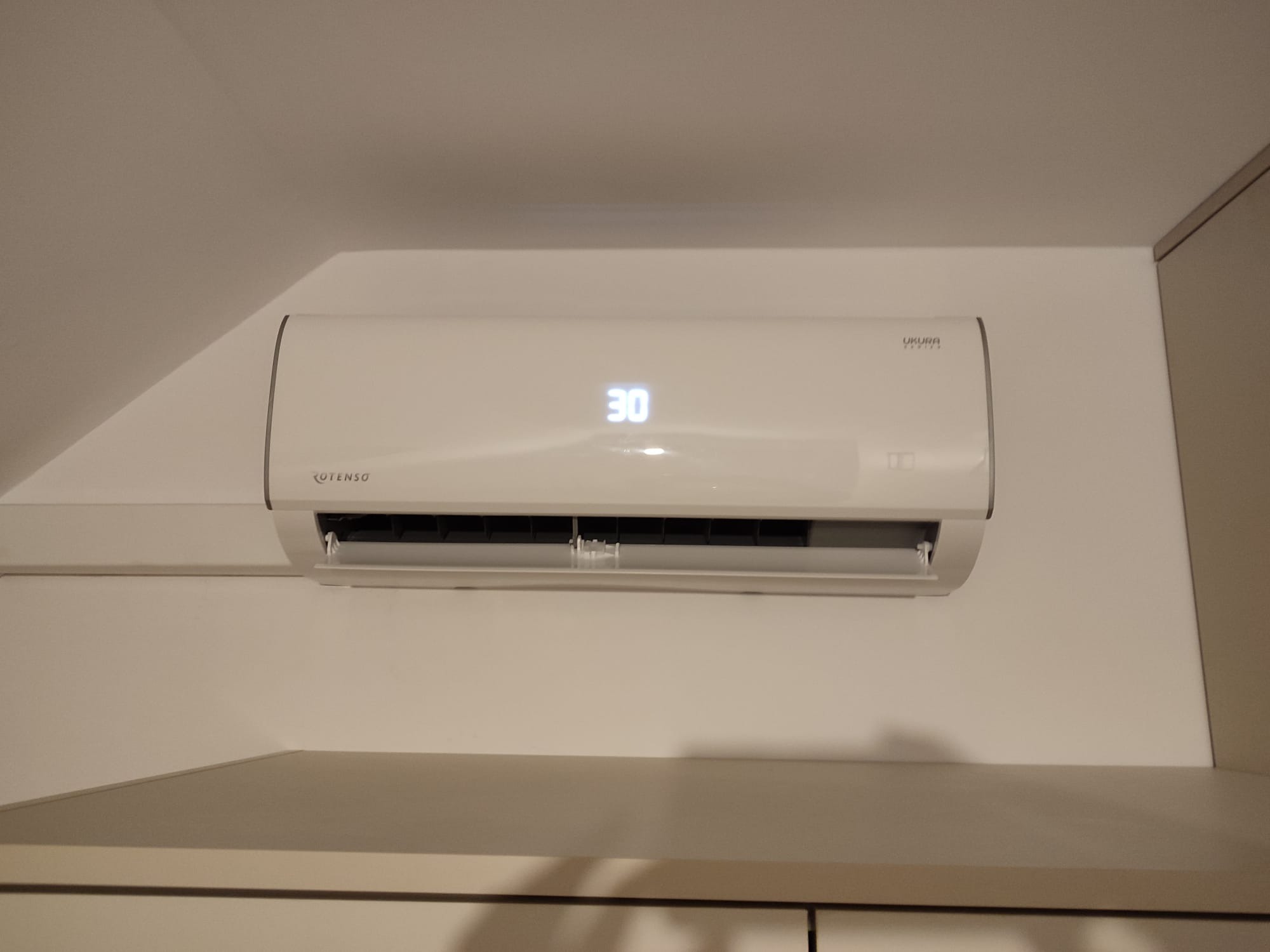 Zamontowany na ścianie, biały klimatyzator marki Rotenso z wyświetlaczem pokazującym temperaturę 30 stopni Celsjusza, w jasnym wnętrzu.