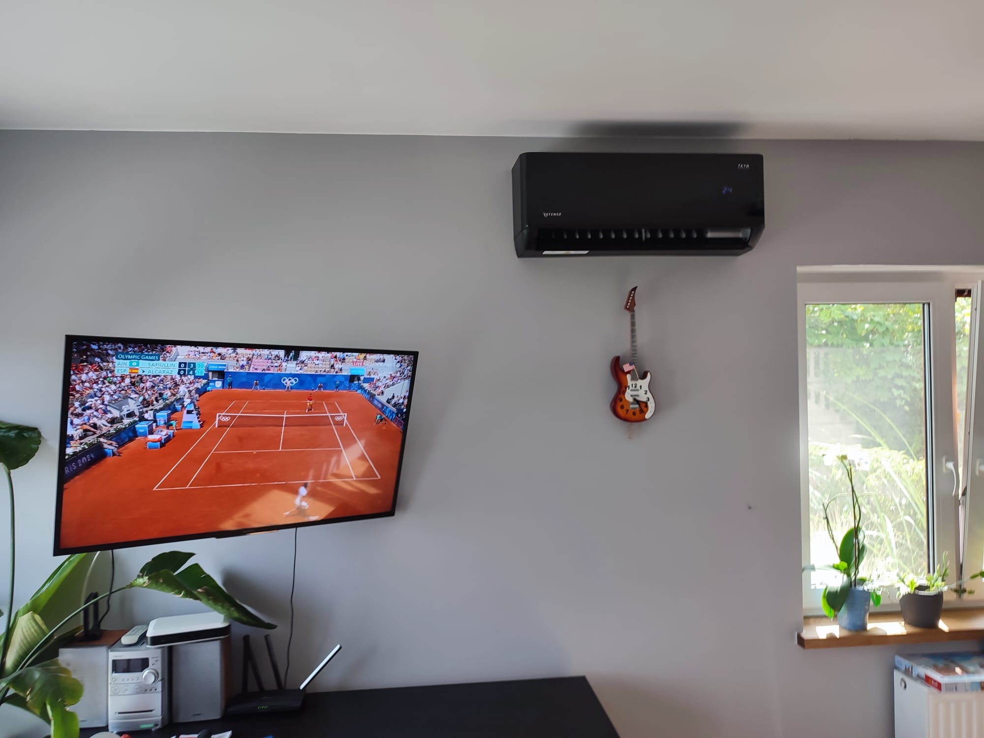 Czarna jednostka klimatyzacyjna zamontowana na szarej ścianie w pokoju, obok telewizora wyświetlającego mecz tenisowy i zegara w kształcie gitary.