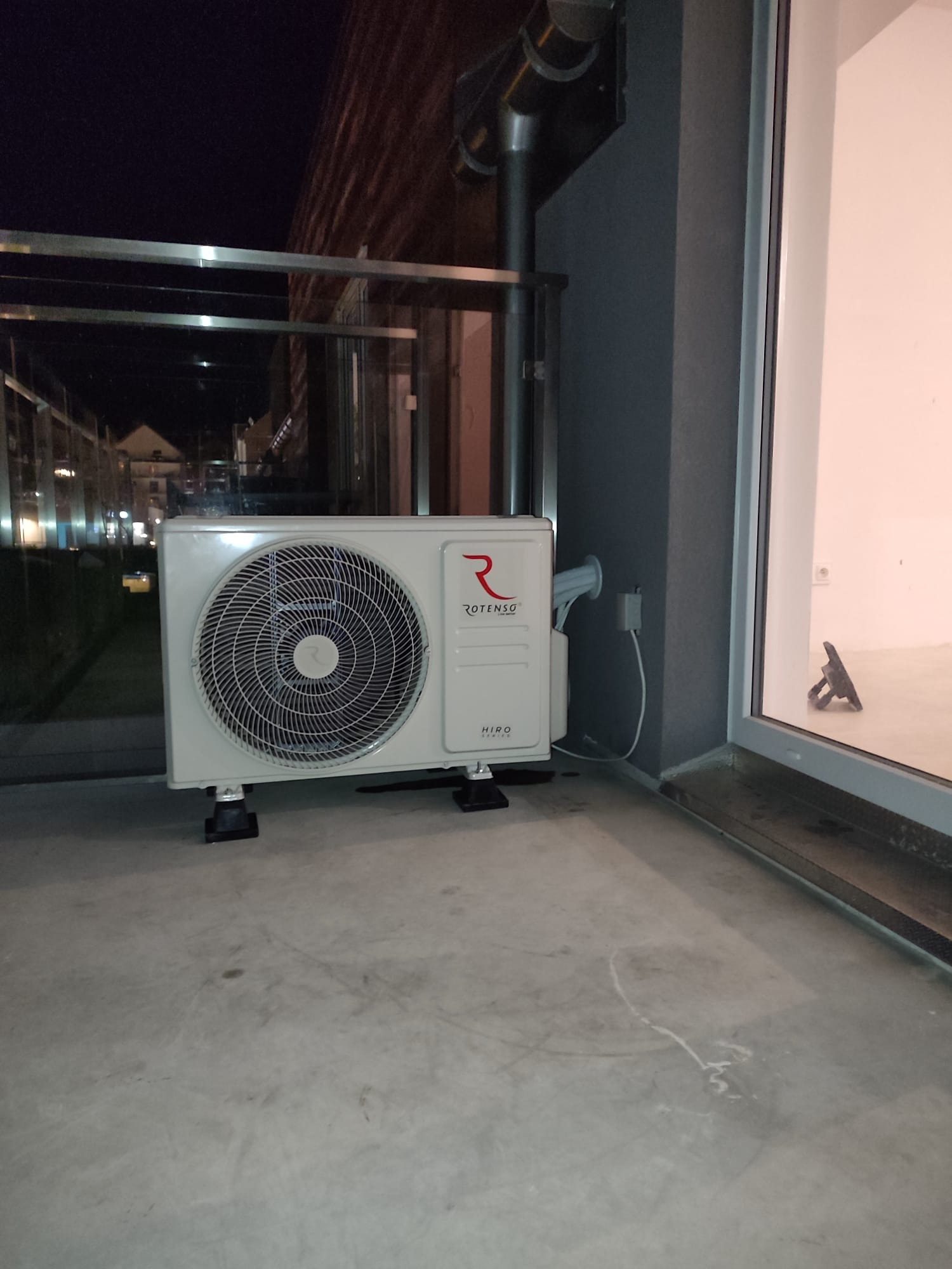 Zewnętrzna jednostka klimatyzacji Rotenso Hiro, zamontowana na balkonie, widoczna instalacja elektryczna i odprowadzająca skropliny. W tle budynki w nocnej scenerii.