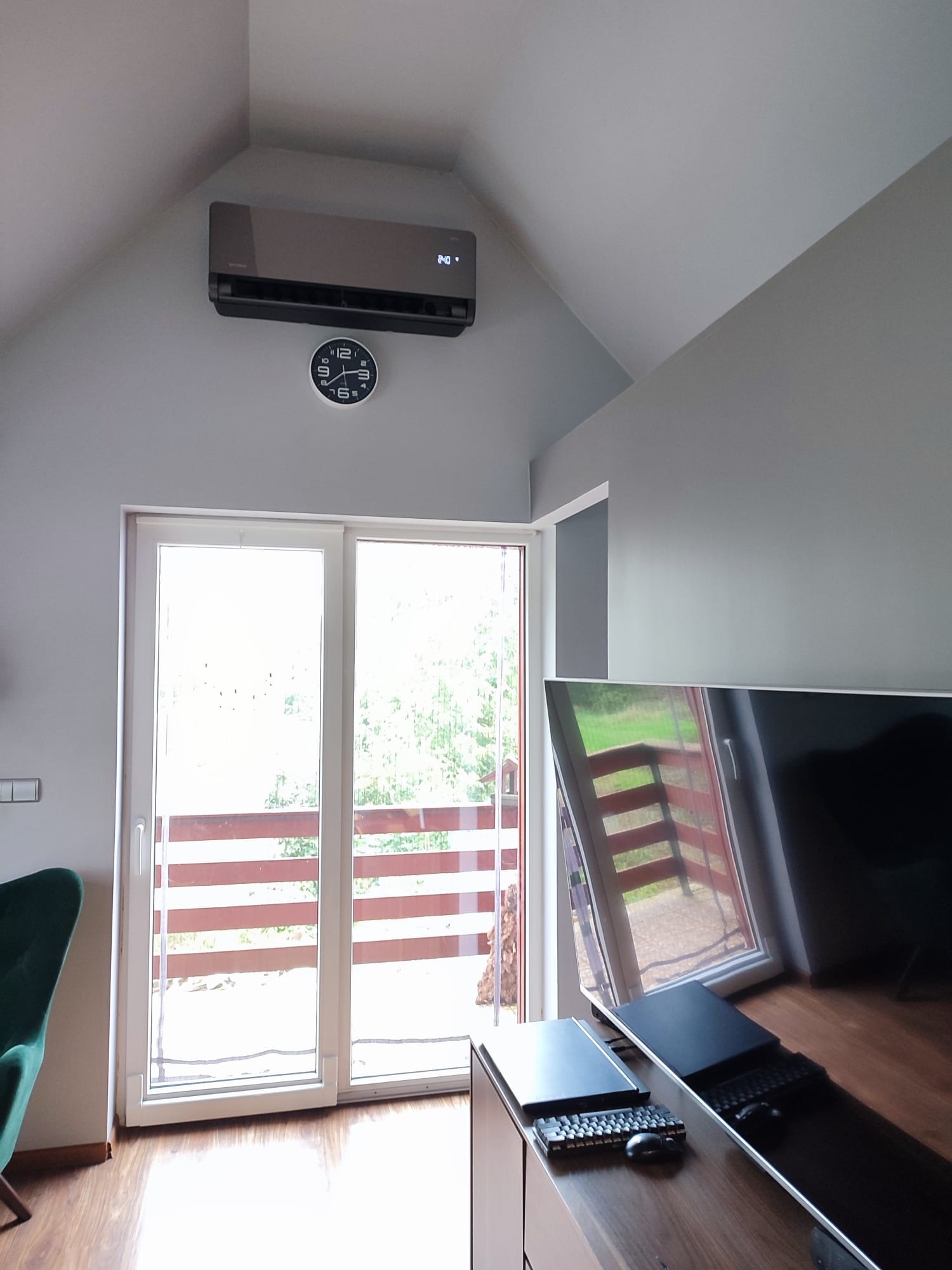 Szary klimatyzator ścienny z wyświetlaczem temperatury, zamontowany w pokoju na poddaszu nad białymi drzwiami balkonowymi, obok okrągłego zegara.