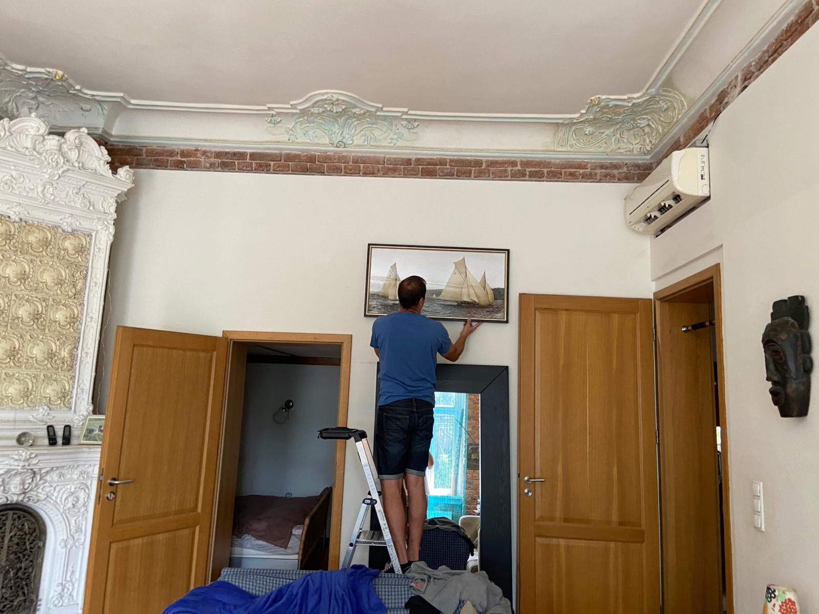 Mężczyzna na drabinie w pomieszczeniu z dekoracyjnym sufitem montuje obraz z żaglówkami, klimatyzator widoczny nad drzwiami po prawej.