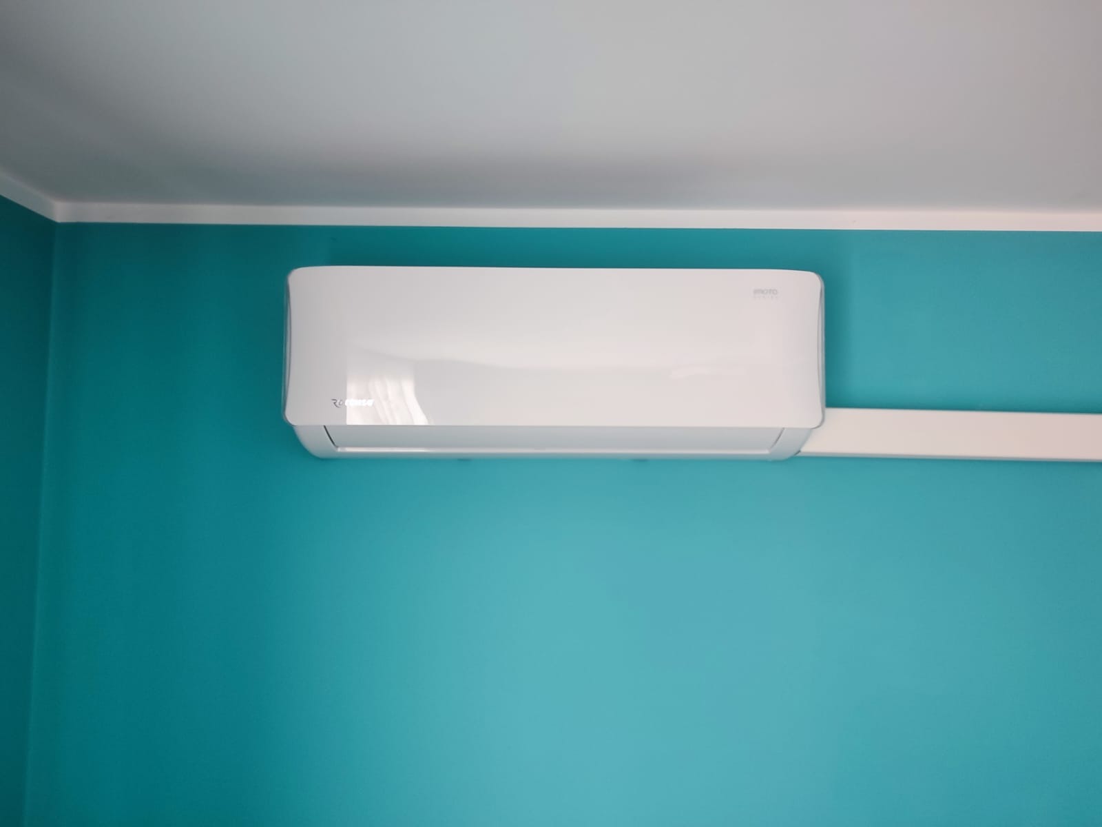 Biały klimatyzator zamontowany na turkusowej ścianie w pomieszczeniu, widoczne przewody w białej listwie.