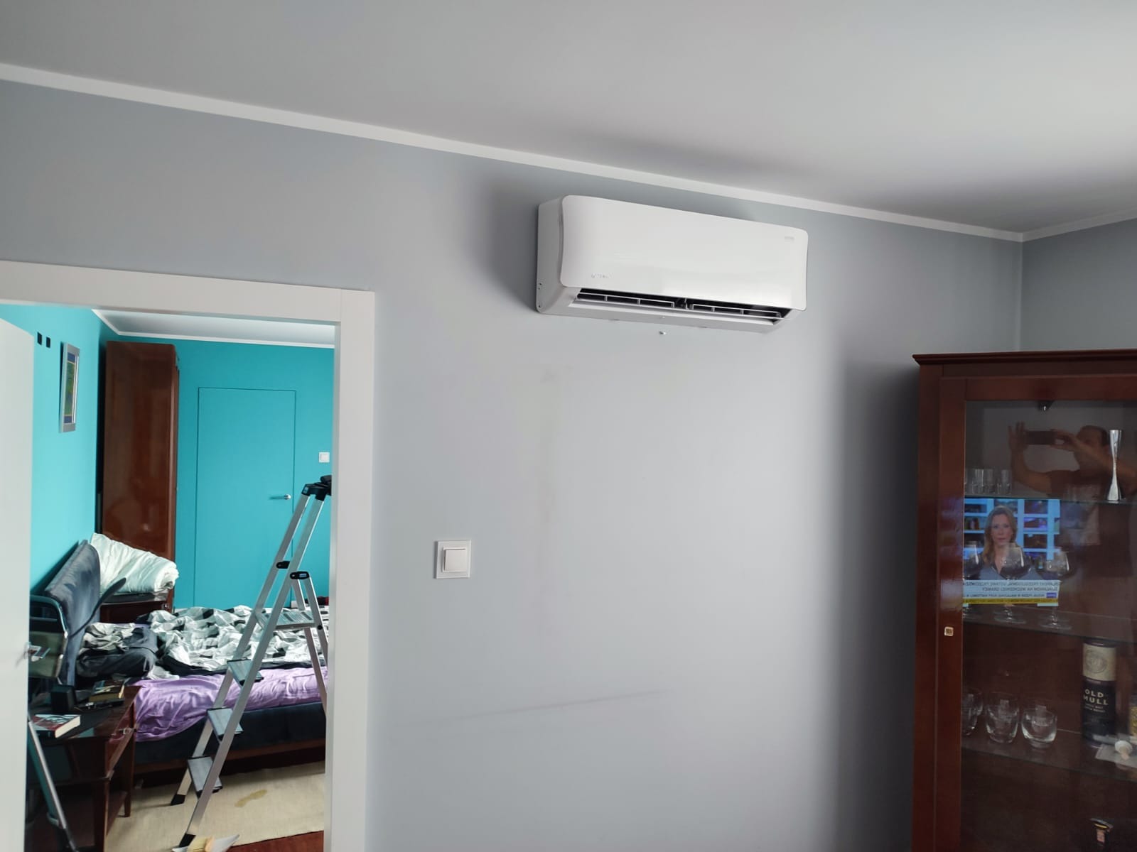 Biały klimatyzator ścienny zamontowany w szarym pokoju, widoczna drabina w przejściu do pokoju z turkusowymi ścianami, obok stoi ciemna witryna.