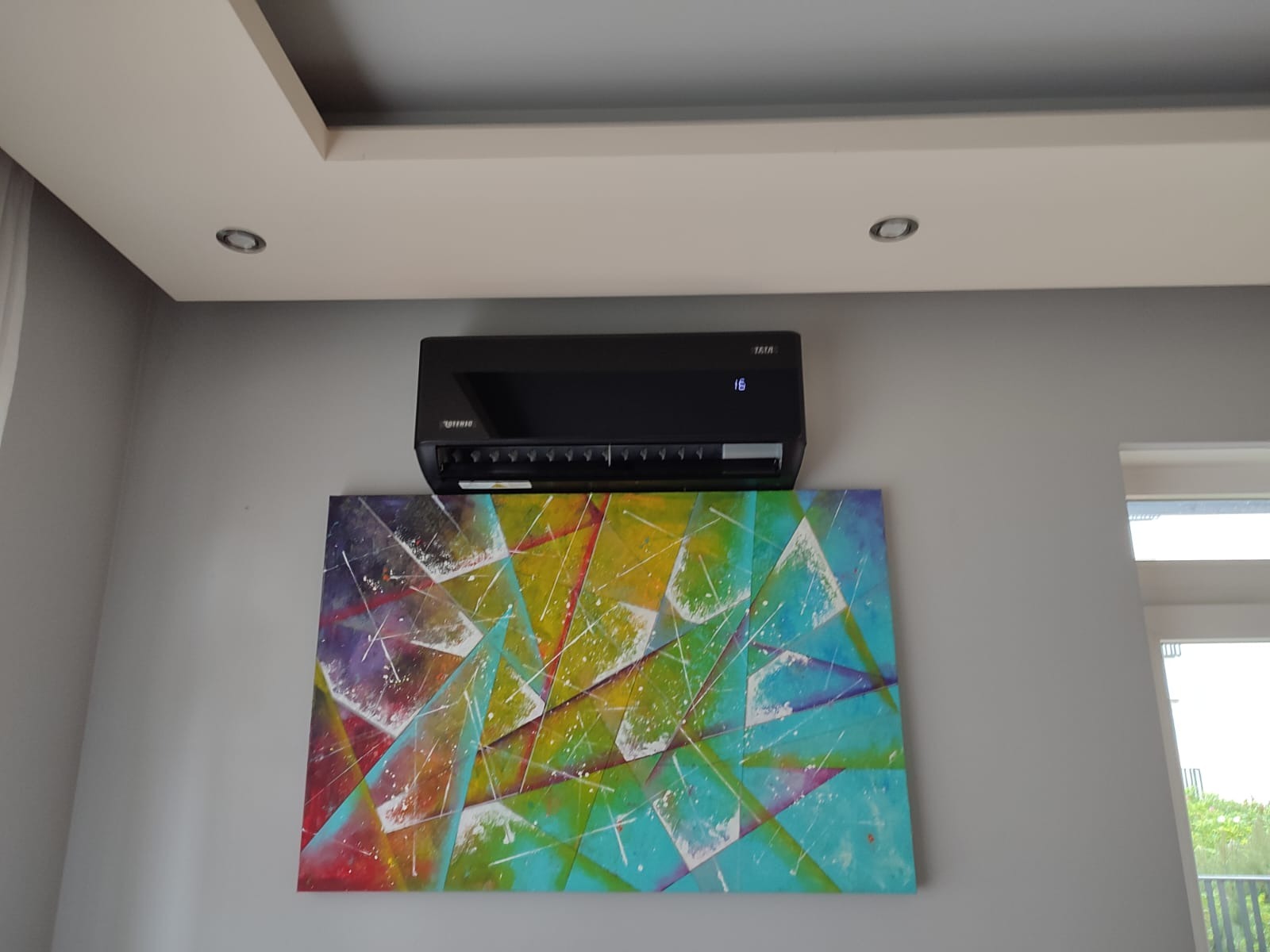 Czarny klimatyzator ścienny z wyświetlaczem temperatury (16 stopni), zamontowany nad abstrakcyjnym, kolorowym obrazem w salonie z szarą ścianą i białym sufitem.