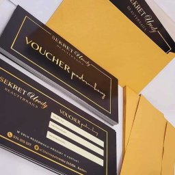 Creative Design Studio - Stos czarno-złotych voucherów podarunkowych z danymi do wypełnienia, obok koperty w kolorze złota, z logo 'Sekret Urody Beauty&Nails'.