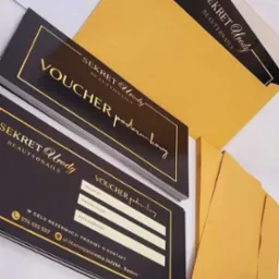 Stos czarno-złotych voucherów podarunkowych z danymi do wypełnienia, obok koperty w kolorze złota, z logo 'Sekret Urody Beauty&Nails'.
