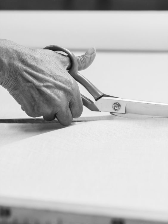 Czarno-białe zdjęcie dłoni krawca tnącej jasną tkaninę dużymi nożycami krawieckimi na stole.