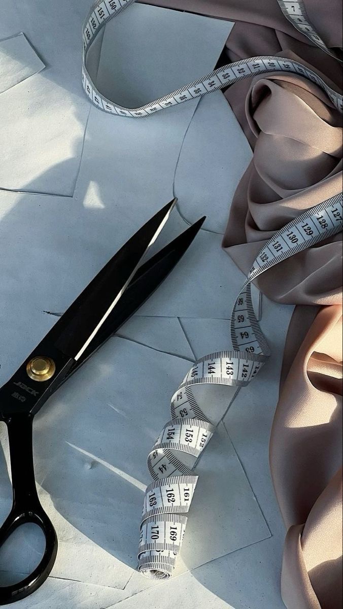 Czarne nożyce krawieckie JACK leżące na białych wykrojach, obok leży zwinięta miara krawiecka i beżowa tkanina.