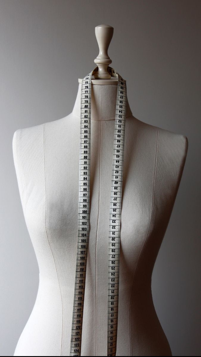 Manekin krawiecki z beżowego płótna z miarką zwisającą wzdłuż tułowia, ustawiony na tle neutralnej ściany.