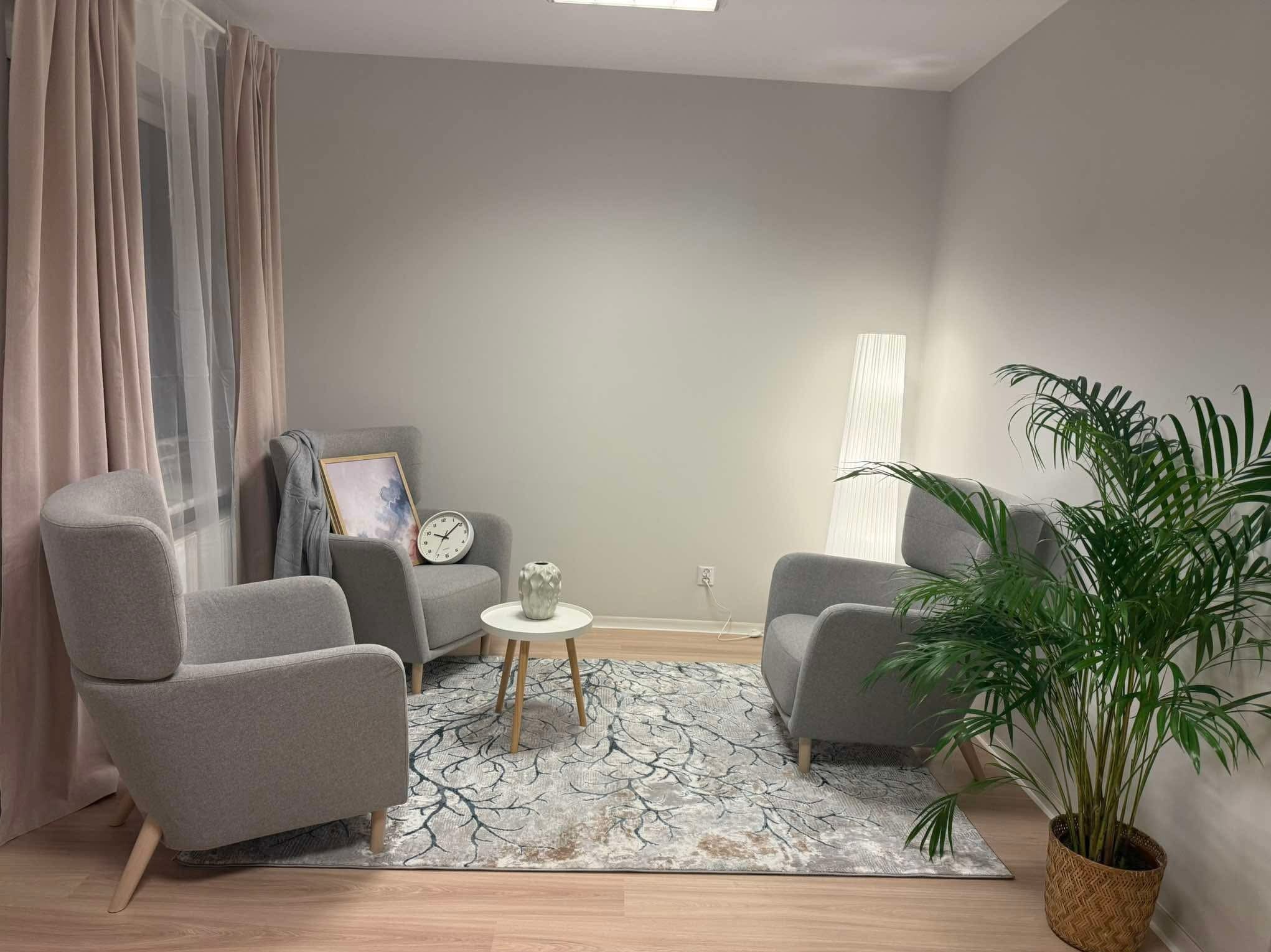 Jasne, minimalistyczne wnętrze gabinetu z trzema szarymi fotelami, stolikiem kawowym z dekoracyjnym wazonem, zegarem i obrazem na ścianie, dywanem z motywem drzewa oraz wysoką lampą i rośliną...