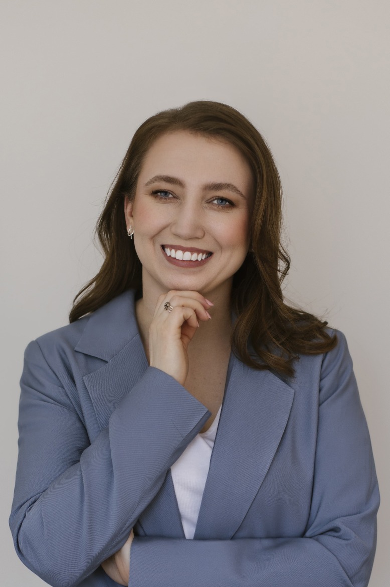 Portret uśmiechniętej kobiety w szarym żakiecie, z ręką podpartą pod brodą, prezentującej pierścionek na palcu.