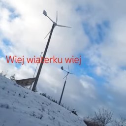 WNES PROSTA SPÓŁKA AKCYJNA - Dwie małe elektrownie wiatrowe na zasypanym śniegiem wzgórzu, pod pochmurnym niebem. Na pierwszym planie widoczny tekst. Ujęcie z dołu.