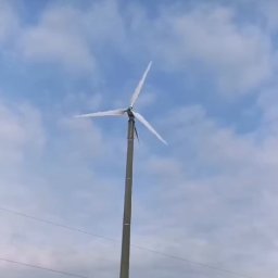 WNES PROSTA SPÓŁKA AKCYJNA - Niska turbina wiatrowa z trzema śmigłami na tle pochmurnego nieba. Widoczne linie energetyczne w dolnej części kadru. Perspektywa od dołu, turbina na metalowym słupie.