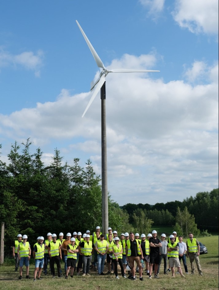 Mikroinstalacja wiatrowa z grupą osób w kaskach i kamizelkach odblaskowych na tle błękitnego nieba z chmurami. Szkolenie dla instalatorów.