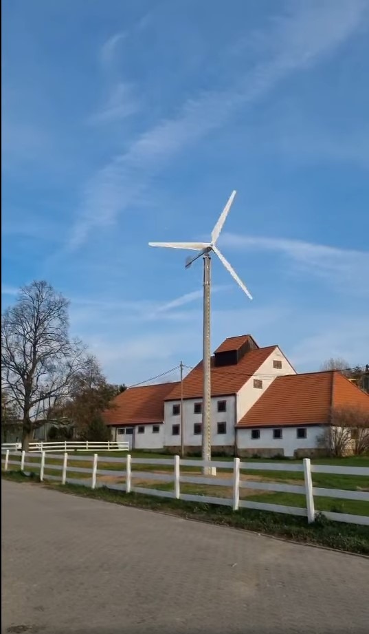 Mała turbina wiatrowa o mocy 15kW, zamontowana na posesji z białym płotem i budynkiem o czerwonym dachu, na tle błękitnego nieba.
