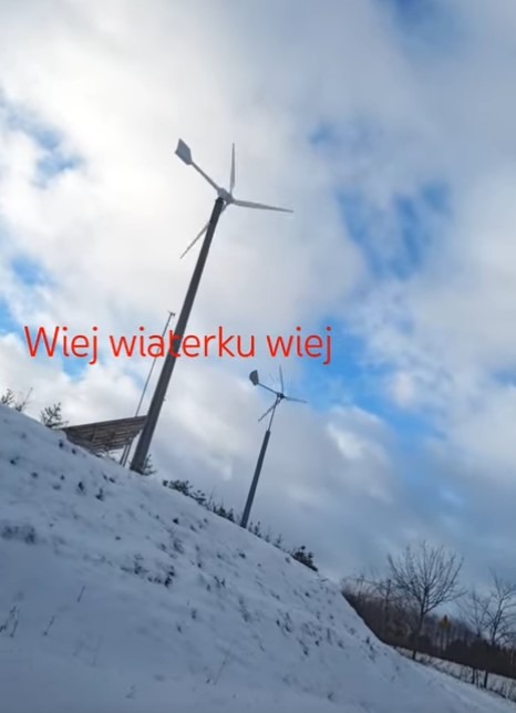 Dwie małe elektrownie wiatrowe na zasypanym śniegiem wzgórzu, pod pochmurnym niebem. Na pierwszym planie widoczny tekst. Ujęcie z dołu.