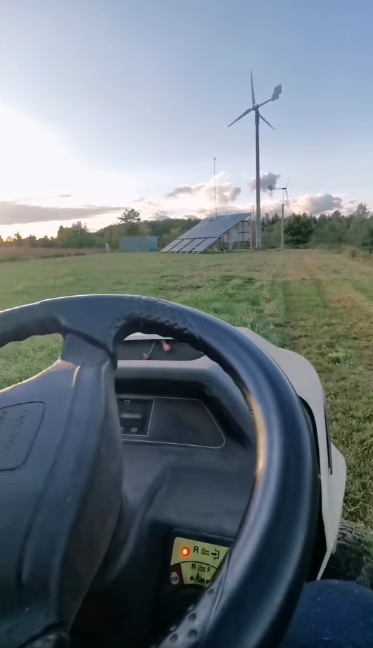 Widok z traktorka koszącego trawę w kierunku turbiny wiatrowej i paneli słonecznych na tle zachodzącego słońca.