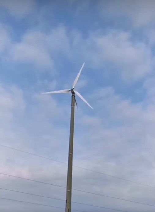 Niska turbina wiatrowa z trzema śmigłami na tle pochmurnego nieba. Widoczne linie energetyczne w dolnej części kadru. Perspektywa od dołu, turbina na metalowym słupie.