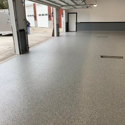 LAKU SPÓŁKA Z OGRANICZONĄ ODPOWIEDZIALNOŚCIĄ - Gładka, szara posadzka żywiczna z delikatnymi płatkami w garażu, widok z otwartej bramy na budynek z białymi bramami garażowymi i czerwoną elewacją, czarne listwy przypodłogowe.