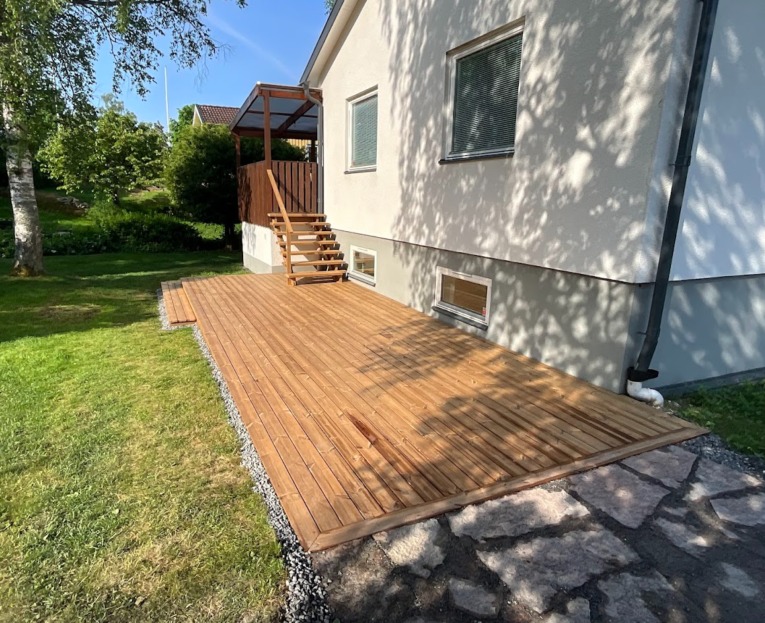 Drewniany taras przylegający do domu z oknami piwnicznymi, wykończony drobnym żwirem od strony trawnika i kamienną ścieżką od strony elewacji.