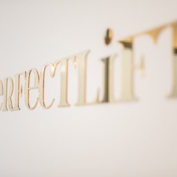 ROBERT JANCZURA PERFECTLIFT - Złoty napis 'perfect lift' na białej ścianie, z efektem rozmycia w tle.