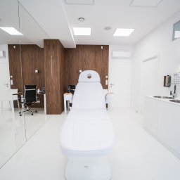 ROBERT JANCZURA PERFECTLIFT - Jasne, minimalistyczne wnętrze gabinetu z białym fotelem zabiegowym na pierwszym planie, widoczne umywalki i dozowniki na ścianie oraz biurko z krzesłem w tle.