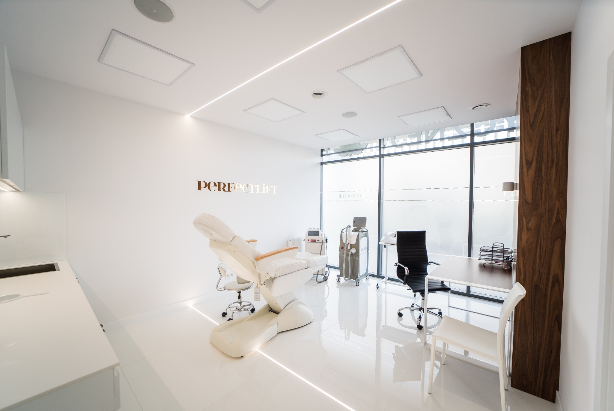 Jasne wnętrze gabinetu z fotelem kosmetycznym, urządzeniami do zabiegów i złotym napisem 'PERFECTlift' na ścianie, minimalistyczny design z białymi ścianami i podłogą.