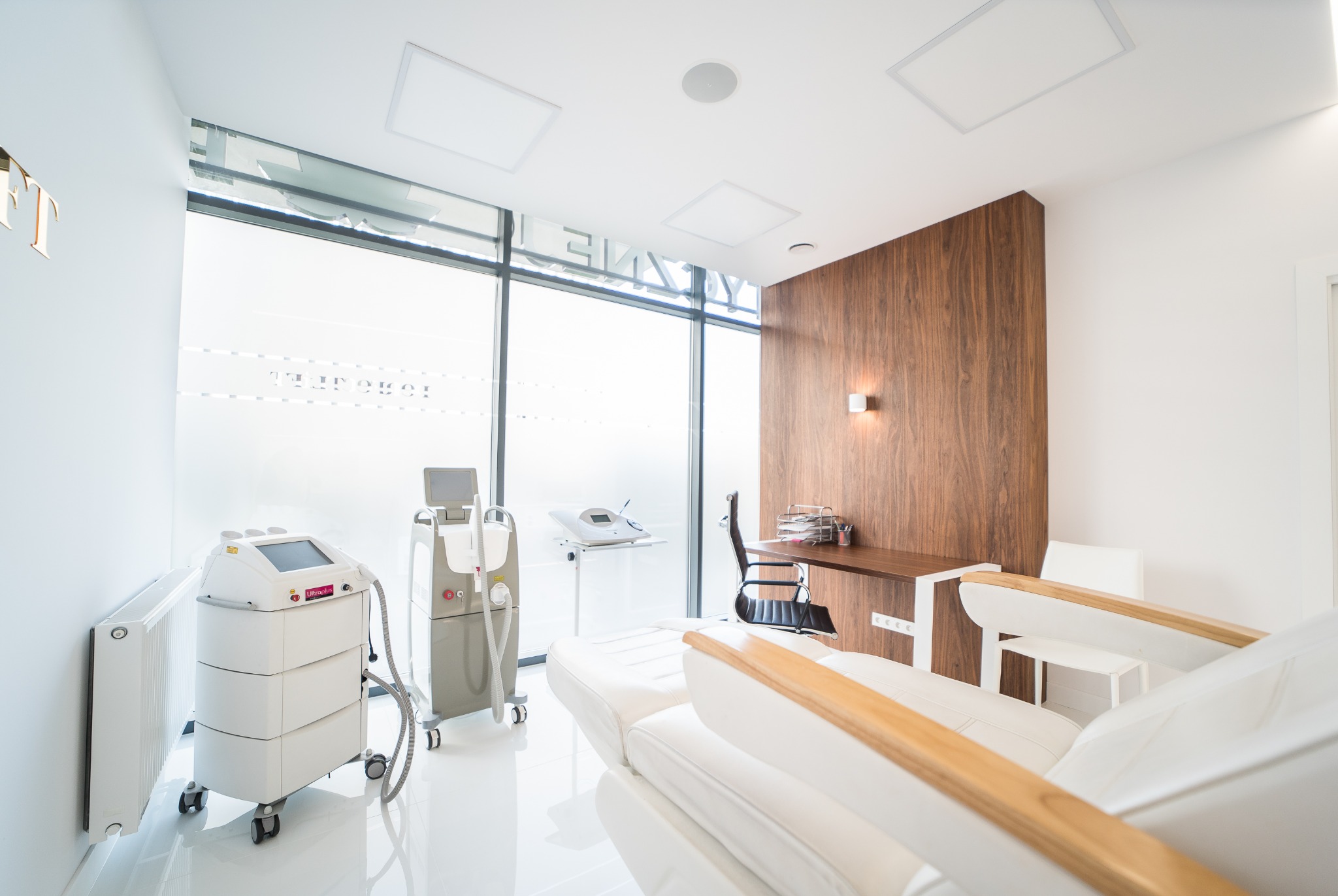 Jasne, minimalistyczne wnętrze gabinetu z białym fotelem zabiegowym, urządzeniami do laseroterapii i biurkiem z drewnianą okładziną ścienną.