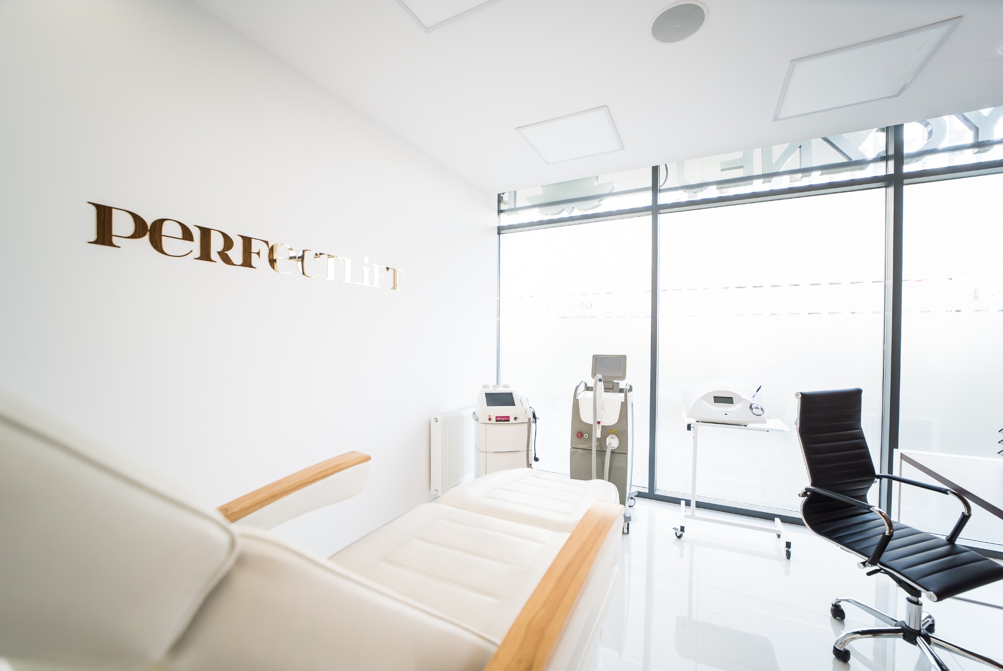 Jasne wnętrze gabinetu z białym fotelem zabiegowym z drewnianymi podłokietnikami, sprzętem kosmetycznym i dużym oknem z widocznym napisem 'Perfect Lift' na ścianie.