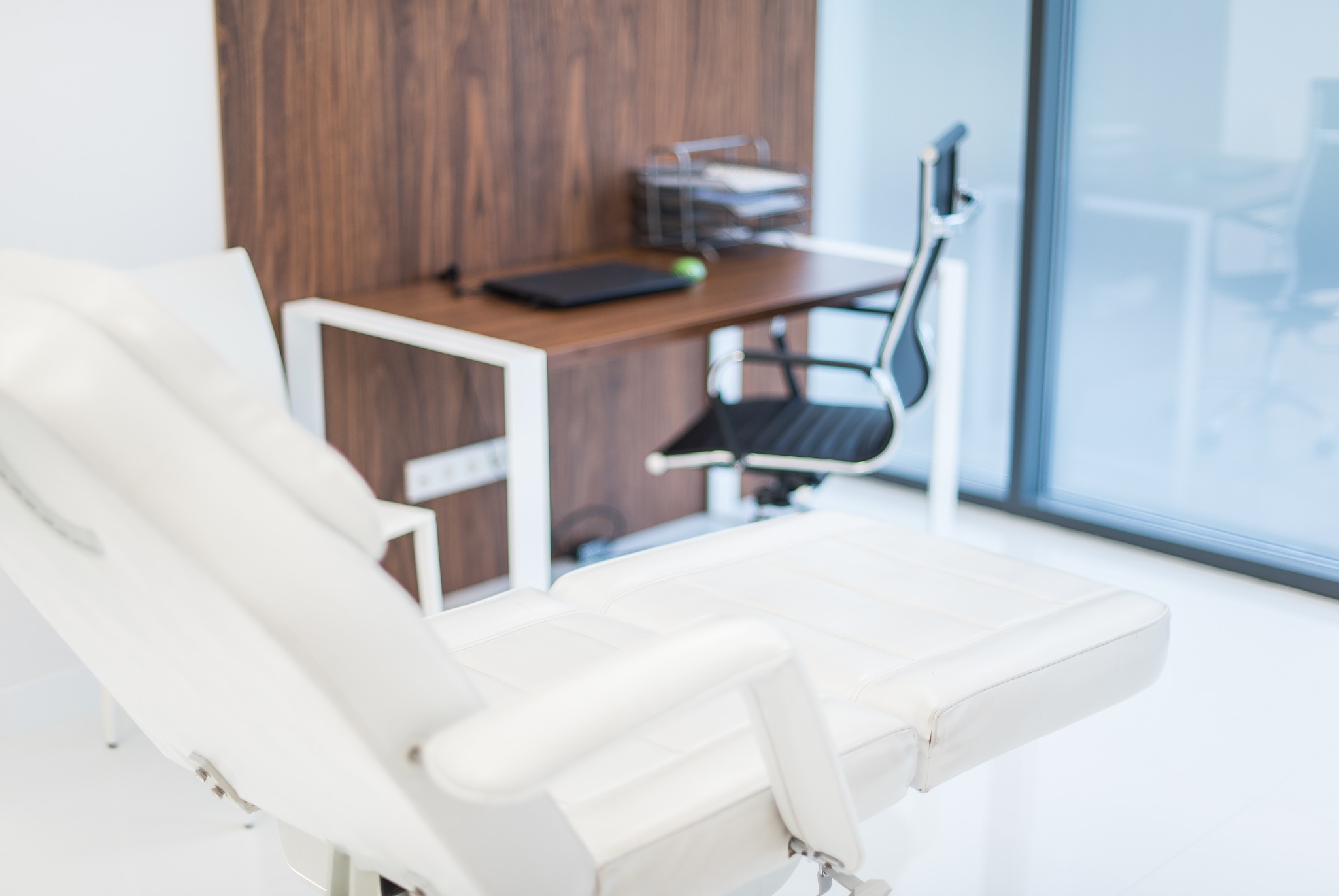 Biały fotel zabiegowy w minimalistycznym gabinecie z biurkiem i krzesłem, widoczne elementy wyposażenia oraz duże okno.