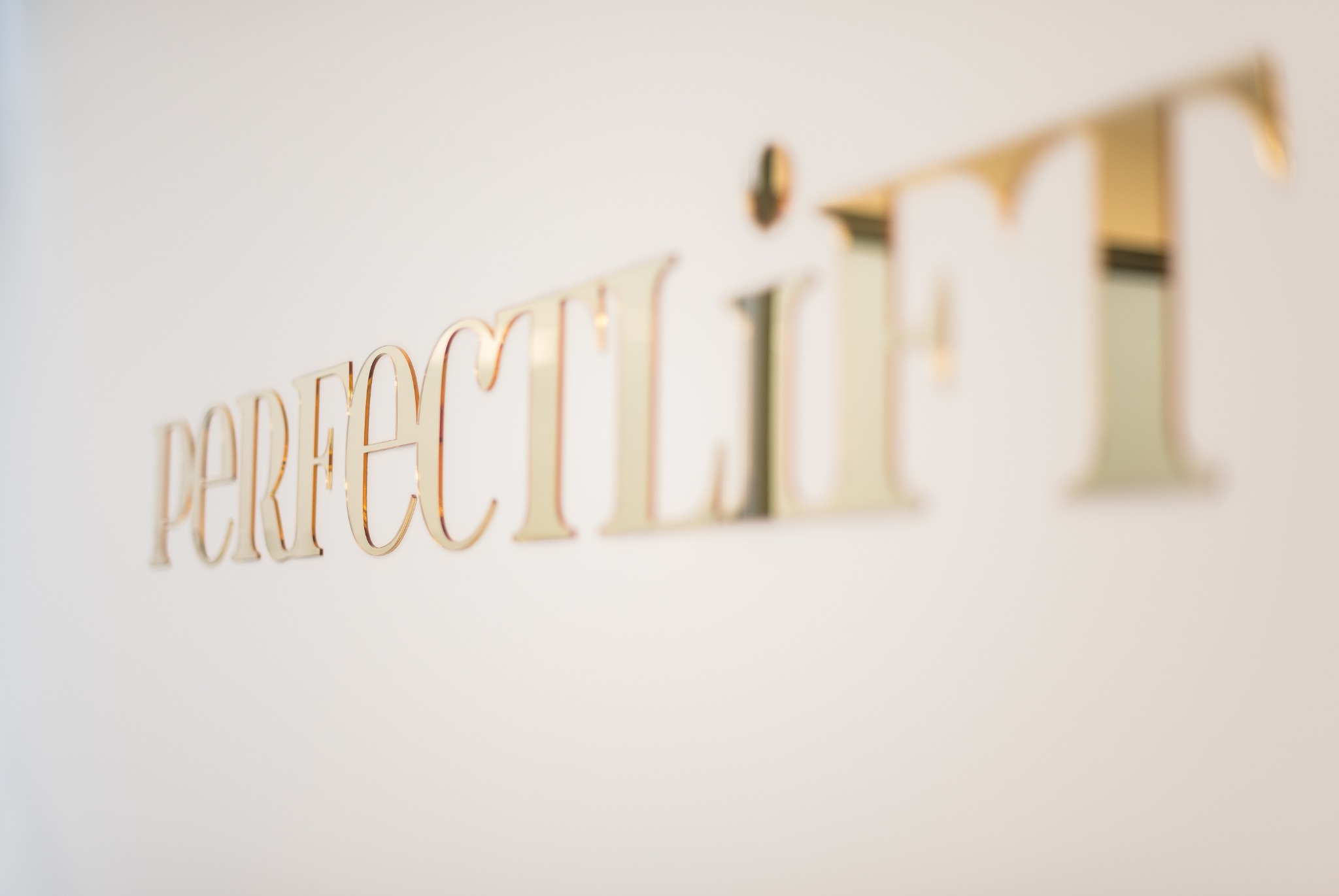 Złoty napis 'perfect lift' na białej ścianie, z efektem rozmycia w tle.