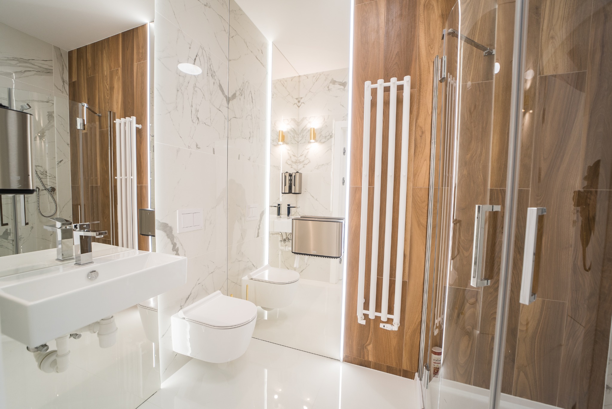 Nowoczesna łazienka z marmurowymi i drewnianymi płytkami, białą umywalką nablatową, grzejnikiem drabinkowym i kabiną prysznicową ze szklanymi drzwiami.