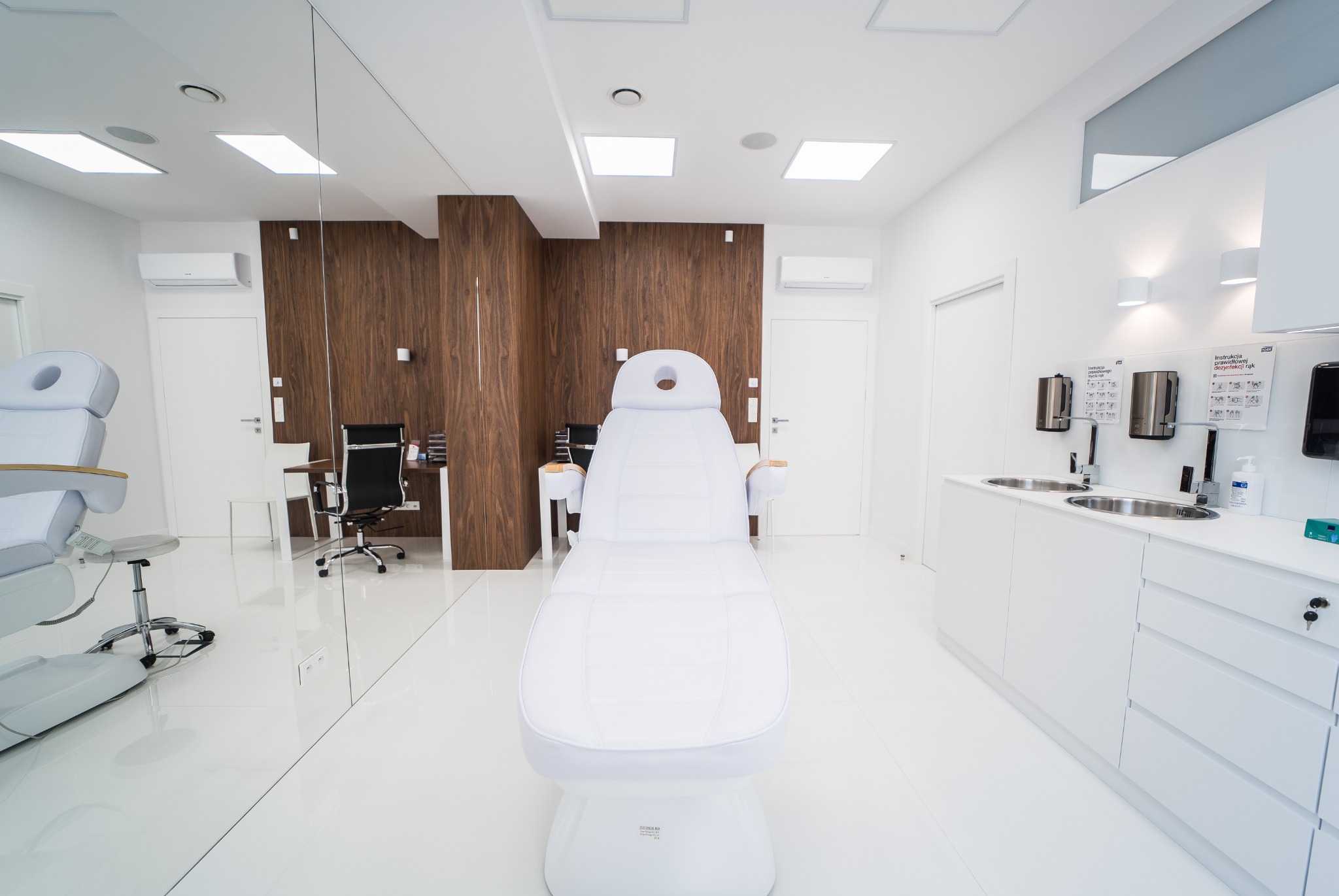 Jasne, minimalistyczne wnętrze gabinetu z białym fotelem zabiegowym na pierwszym planie, widoczne umywalki i dozowniki na ścianie oraz biurko z krzesłem w tle.
