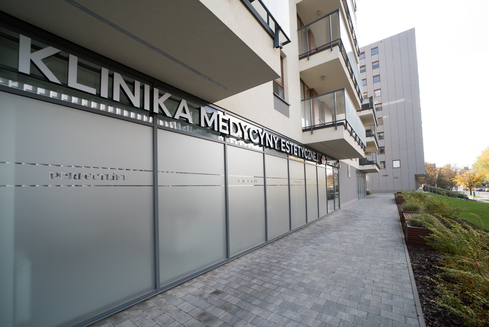 Fasada nowoczesnej kliniki medycyny estetycznej z napisem 'KLINIKA MEDYCYNY ESTETYCZNEJ' nad matowymi oknami i nazwą 'Perfect Lift' na szklanych panelach, widok z chodnika.