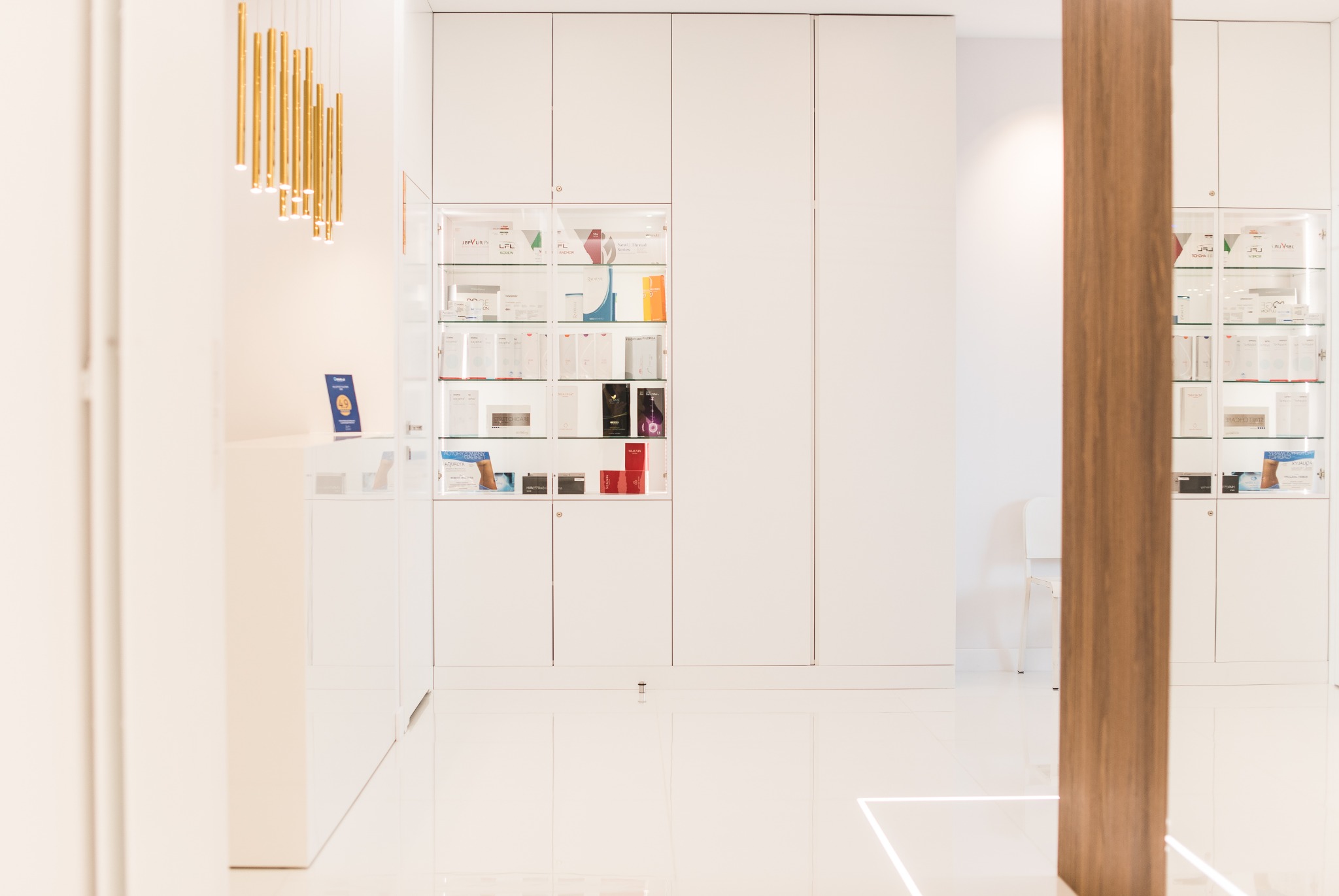 Jasne wnętrze gabinetu z białymi szafkami ekspozycyjnymi wypełnionymi kosmetykami i preparatami, oświetlone złotymi lampami wiszącymi.
